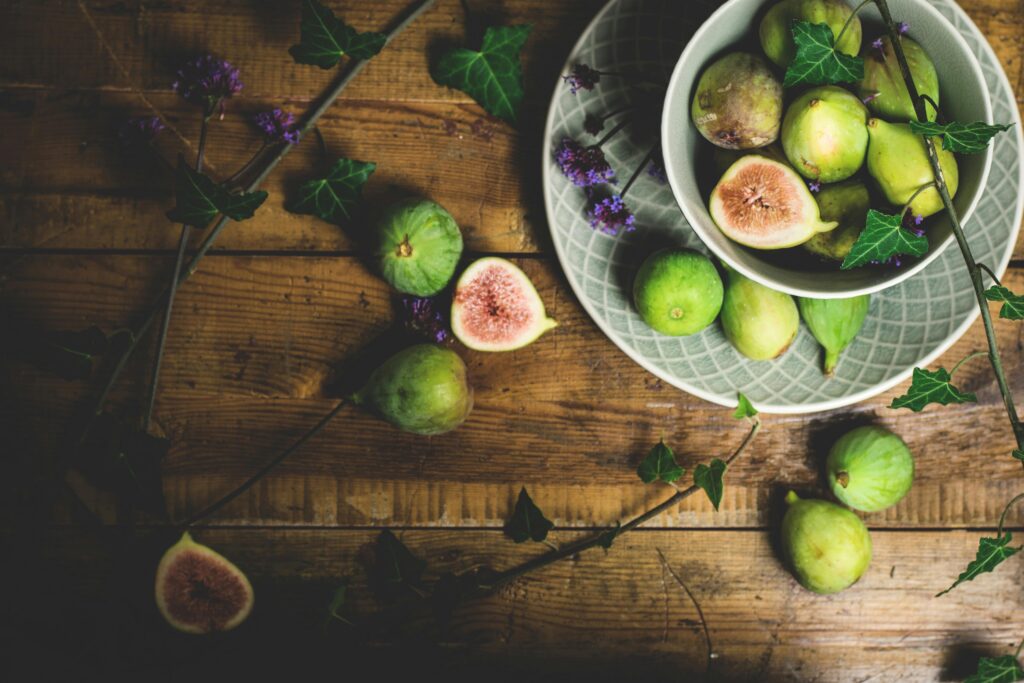 A rustic flat lay of fresh green figs in a bowl on a wooden table, complemented by ivy leaves.
